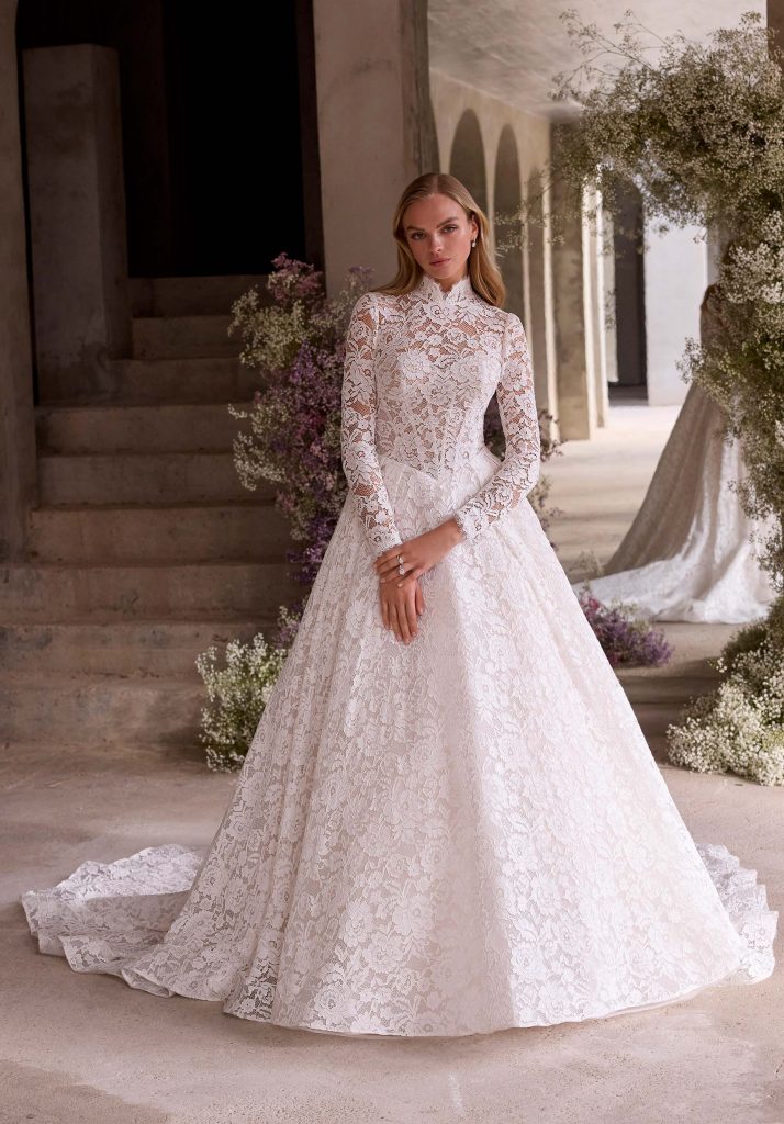 A bride in a high-neck lace long sleeve wedding dress with a voluminous skirt, standing near stone steps and flowers indoors.