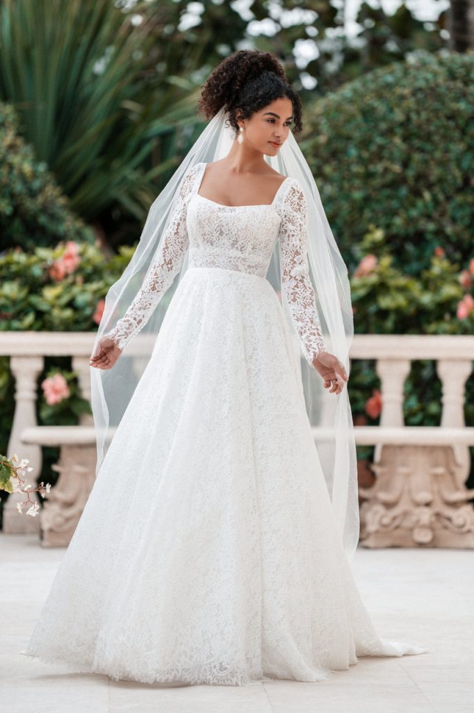 A bride in a square neckline lace long sleeve wedding dress with a full skirt and veil, standing outdoors near greenery and flowers.