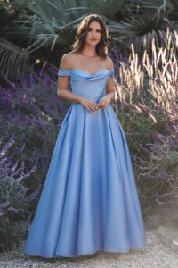 Woman in a light blue off-shoulder evening gown standing in front of lush greenery.