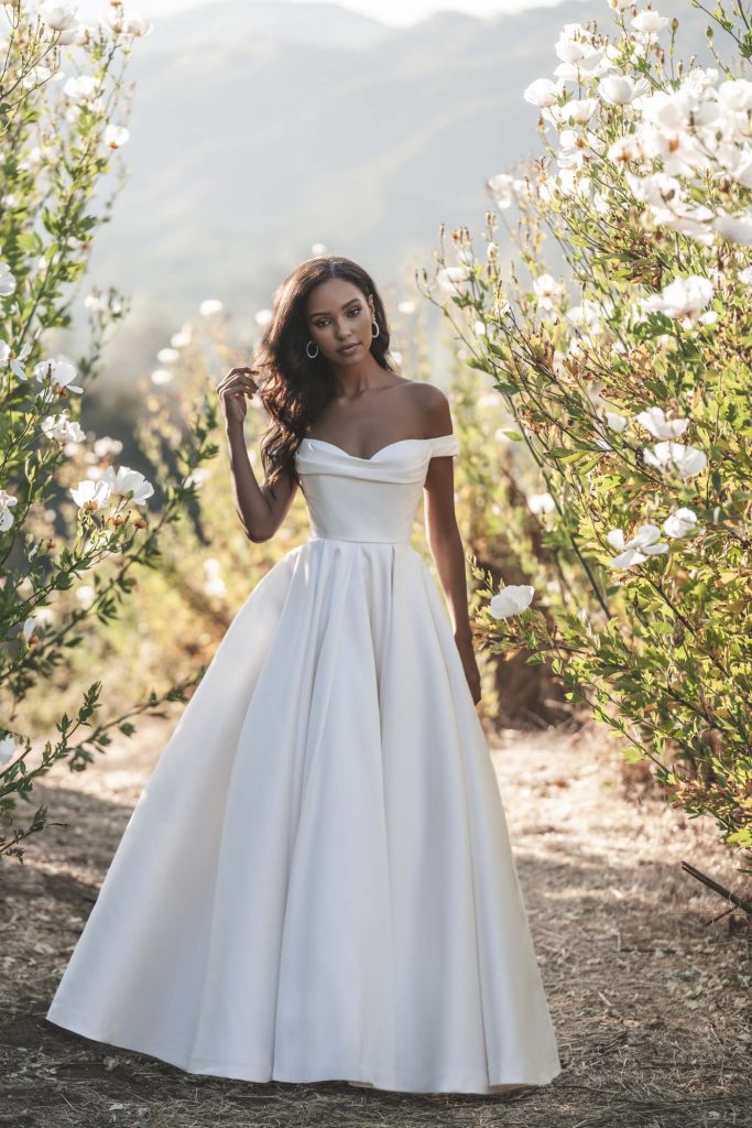 A bride stands among tall white flowers wearing an off-the-shoulder satin ball gown with a pleated skirt.