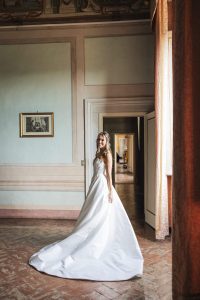 Bride wearing a sleek satin wedding gown with a long train posing in an elegant vintage-style room.