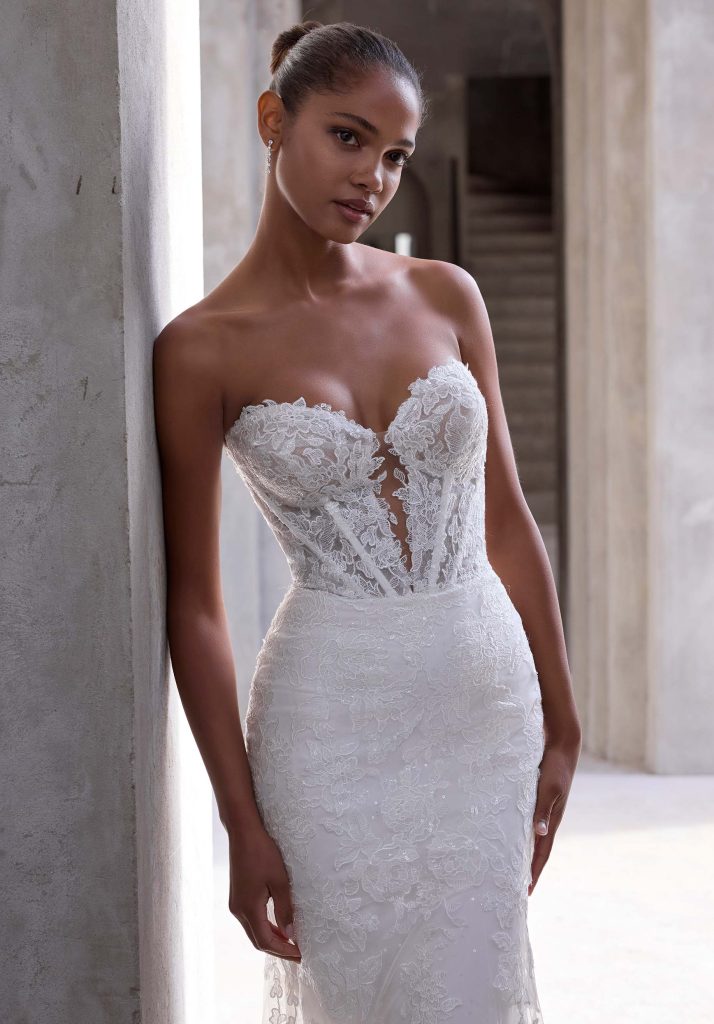 A woman in a strapless lace wedding gown with a plunging neckline and fitted silhouette leans against a stone column, looking at the camera with a calm expression.