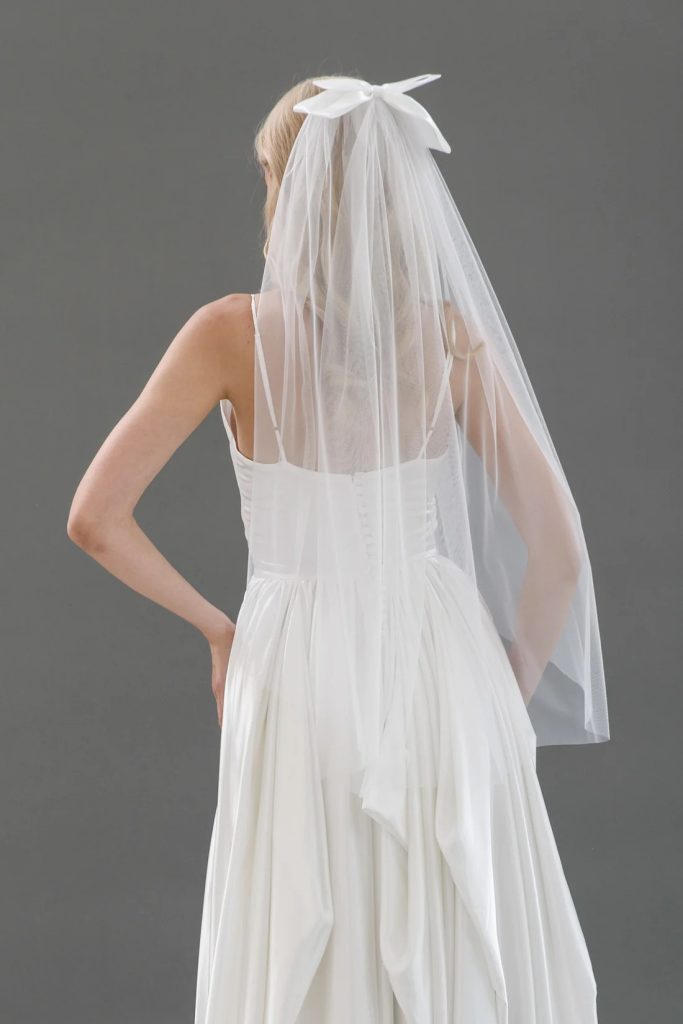 A bride in a white gown is shown from the back wearing a sheer veil tied with a simple white bow at the crown of her head.
