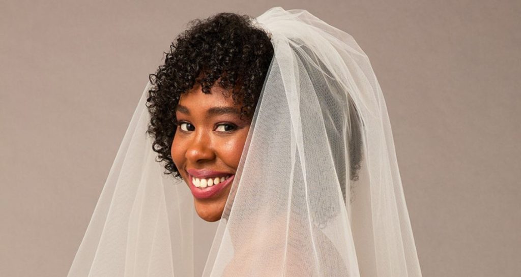 A bride smiles while wearing a layered veil over her curly hair, shown from the shoulders up against a neutral background.
