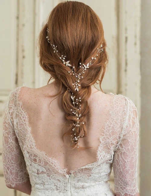 A bride with long red hair styled in a loose braid wears a delicate vine-like hair accessory intertwined throughout the braid, shown from the back.