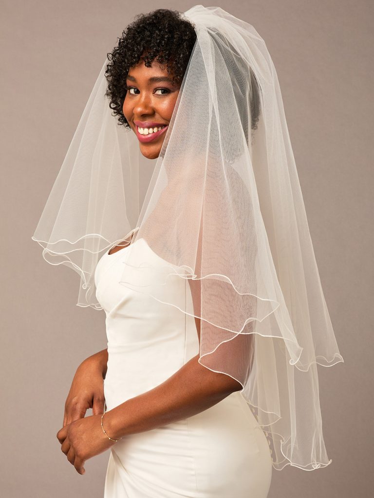 A bride wearing a fingertip-length layered veil smiles while standing in profile in a simple white gown against a neutral backdrop.
