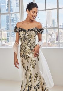 A woman wearing an off-the-shoulder gold gown with floral detailing smiles near large windows overlooking a city skyline.