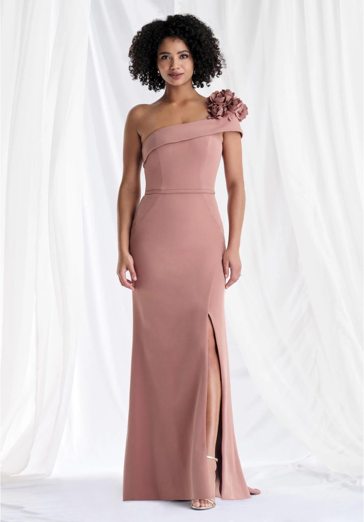 A woman wearing a blush one-shoulder gown with a thigh-high slit stands in front of soft white draping.