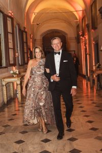 A smiling couple walks arm in arm through a grand hallway, the woman in a patterned one-shoulder gown and the man in a black tuxedo.