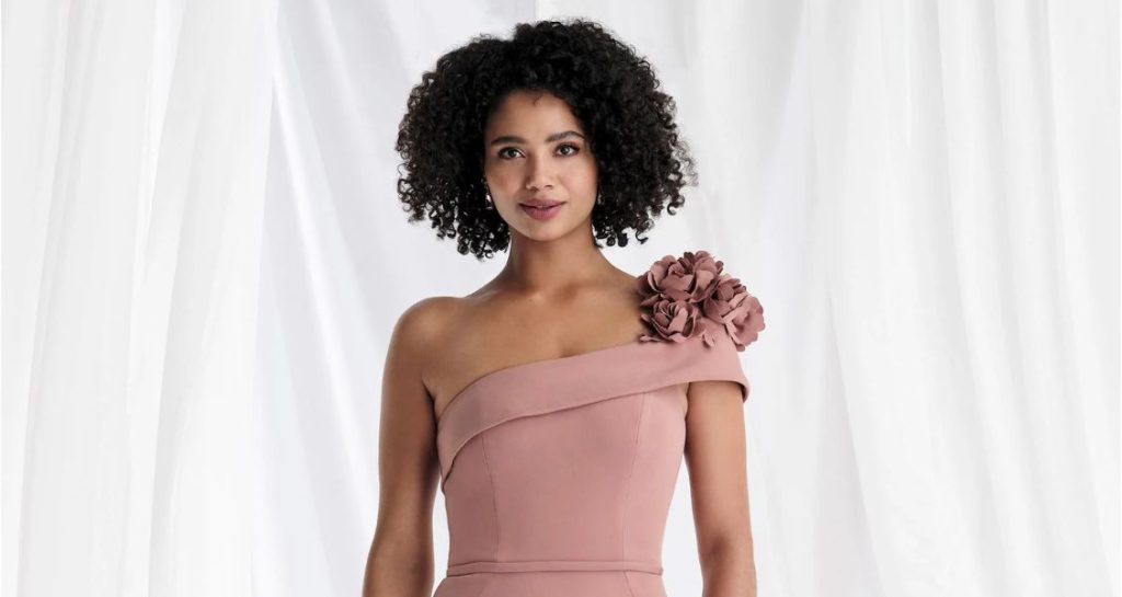 A woman in a blush one-shoulder gown with large floral appliqués on the strap stands against a white draped backdrop.