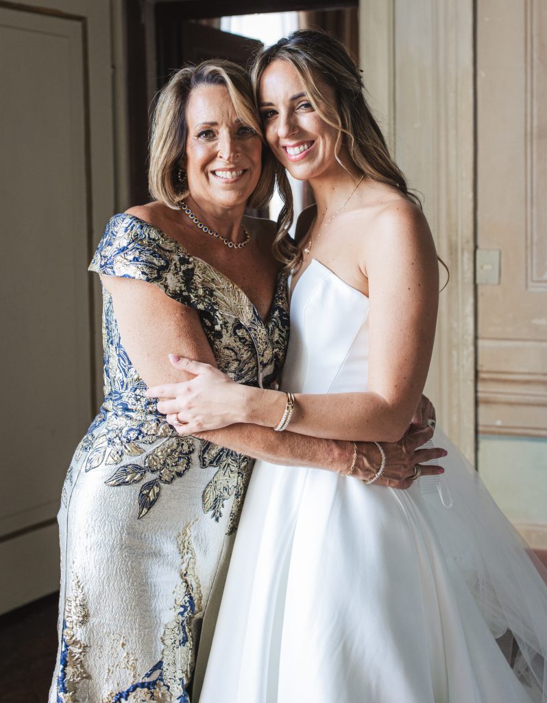 A woman in a gold and navy gown poses with a smiling bride in a white dress and long veil in an ornate room.
