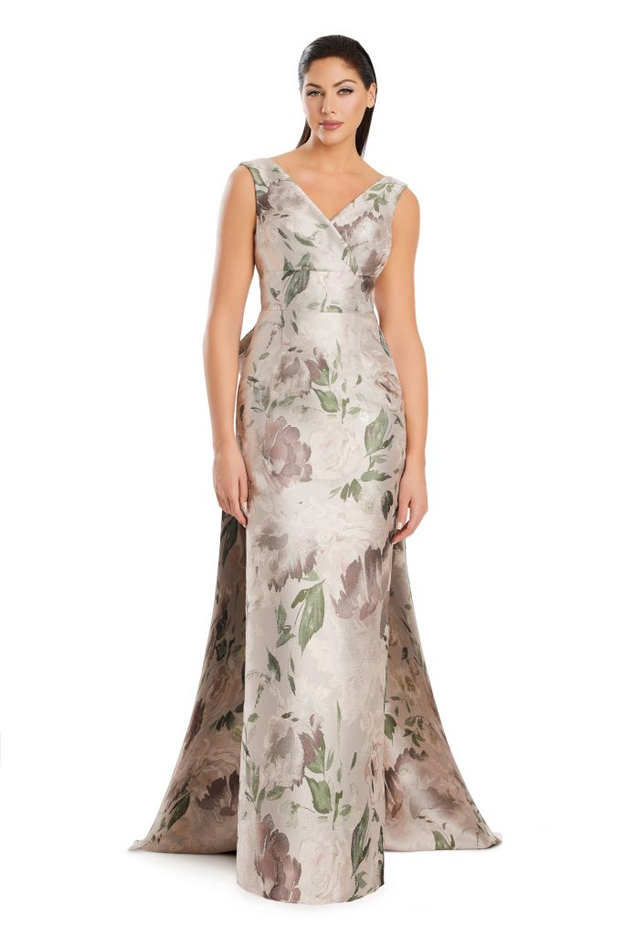 A woman wearing a fitted floral brocade gown with a dramatic train stands facing forward against a white background.