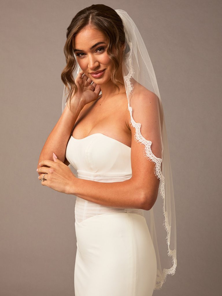 A bride in a strapless white gown touching her hair while wearing a fingertip-length veil with lace edging.