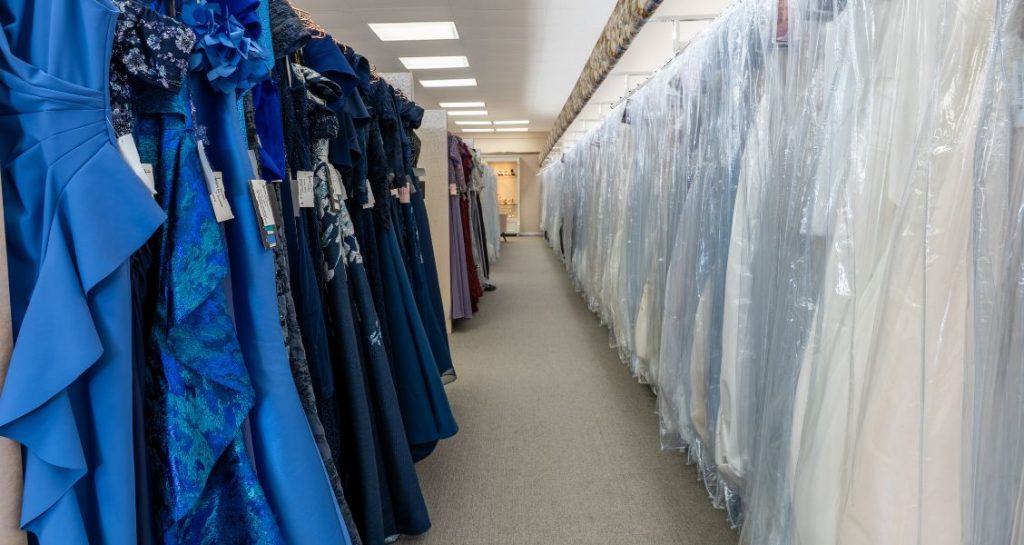 Rows of blue evening gowns hang on one side of an aisle while wedding dresses covered in protective plastic line the opposite side inside a bridal shop.