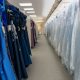 Rows of blue evening gowns hang on one side of an aisle while wedding dresses covered in protective plastic line the opposite side inside a bridal shop.