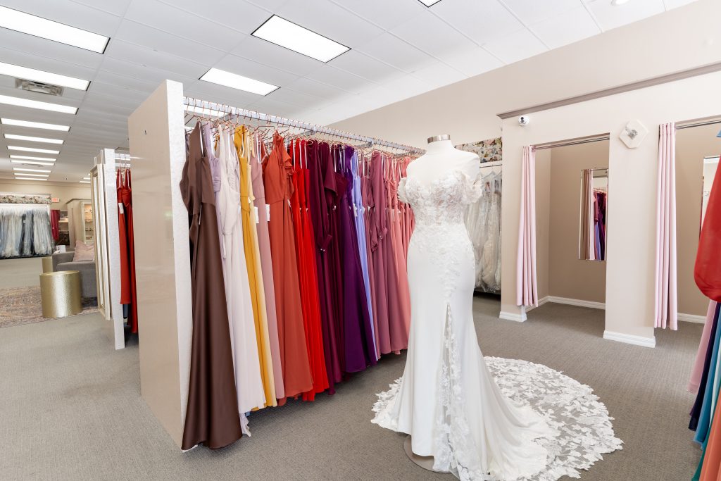 A white lace wedding gown with an off-the-shoulder neckline is displayed on a mannequin next to racks of bridesmaid dresses arranged by color inside a bright bridal shop.