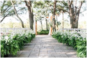 An outdoor wedding ceremony aisle is lined with lush greenery and white flowers leading to a floral arch beneath tall trees.
