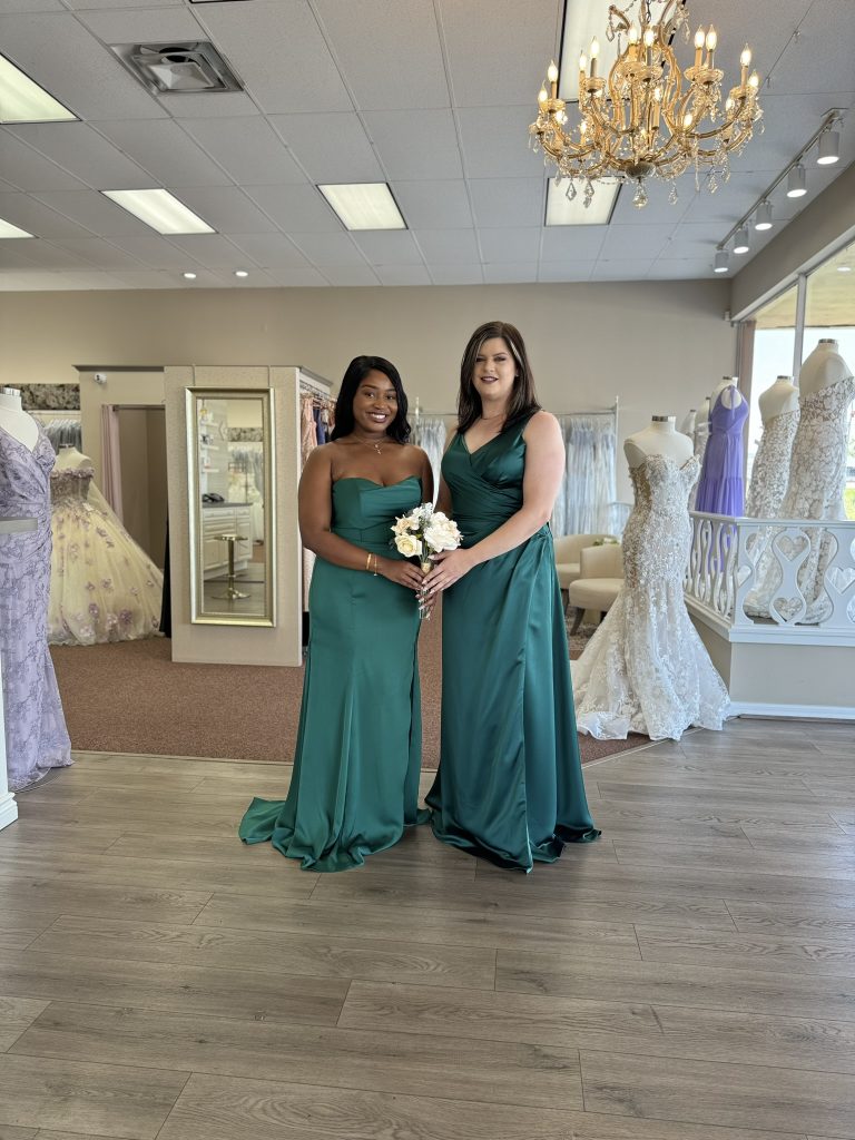 Two women wearing matching emerald green bridesmaid dresses stand together inside a bridal shop, holding a small bouquet and smiling at the camera.