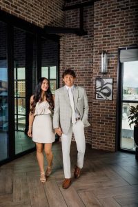 A man and woman in a grey suit and beige dress walk through the interior of a brick building, smiling and holding hands.