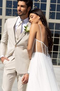 A bride leans her head on the groom’s shoulder, wearing a white gown with a sheer pearl-detailed back, while the groom stands beside her in a light beige suit holding a white rose boutonniere.