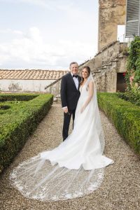 A bride and an older man stand together on a gravel path near stone buildings, with the bride wearing a long white gown and veil.