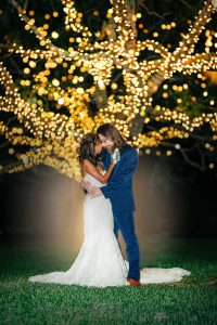 A bride and groom embrace at night under glowing string lights in an outdoor setting.