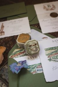 A flat-lay arrangement shows wedding invitation pieces, green envelopes, and two rings placed inside a velvet ring box.