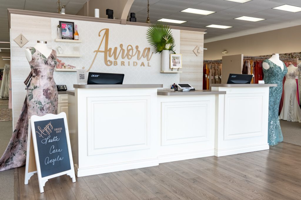 A bridal shop reception area with white counters, mannequins in formal dresses, and a floral-accent wall behind the front desk.