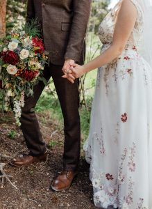 A bride in a floral embroidered gown holds hands with a groom wearing a brown suit, shown from the shoulders down, with the bride holding a large bouquet of red, white, and green flowers.