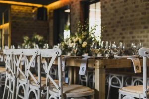 A long wooden dining table is set with glassware, neutral linens, and floral arrangements inside a warmly lit brick reception space.