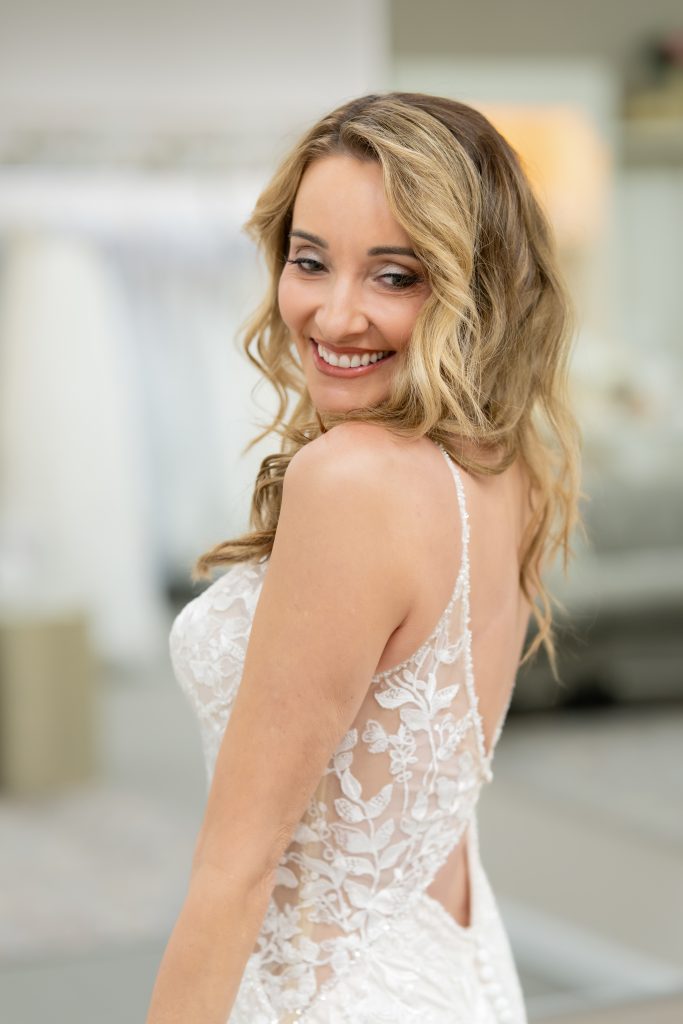 A close-up of a bride smiling over her shoulder, showing the lace back detail of her wedding dress.