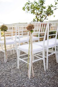 A row of white wooden chairs lined up outdoors on gravel, each decorated with a single hydrangea bloom and long pastel ribbons hanging down the side.