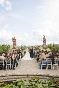 A bride and groom stand together at an outdoor ceremony altar with seated guests on both sides in a scenic garden setting.