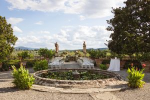 An outdoor wedding ceremony setup featuring white chairs arranged in rows, large stone statues, lush greenery, and a circular pond in the foreground under a bright sky.