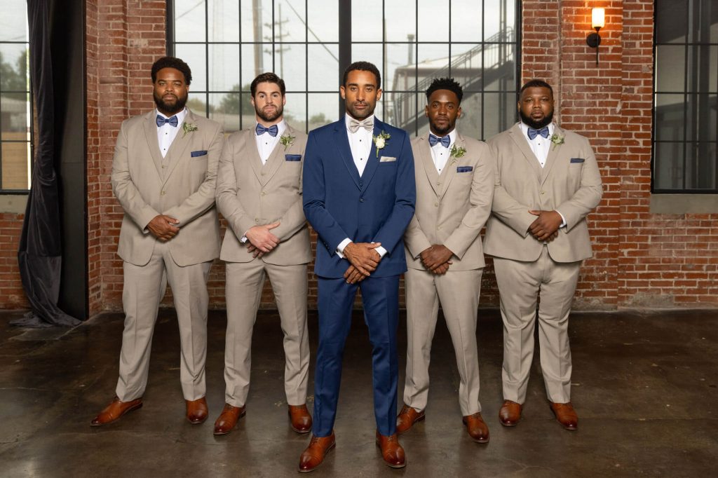 Five groomsmen stand in a line in front of a large industrial window, with the groom in a blue suit at the center and the other men wearing matching tan suits with blue bow ties.