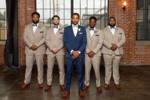 Five groomsmen stand in a line in front of a large industrial window, with the groom in a blue suit at the center and the other men wearing matching tan suits with blue bow ties.