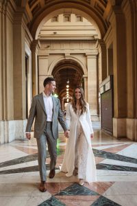 A bride and groom walk hand in hand through an arched hallway, with the bride wearing a lace jumpsuit layered under a long white coat.