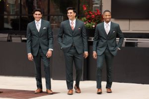 Three groomsmen walk side by side outdoors wearing matching dark green suits, white shirts, and coordinating ties while smiling.