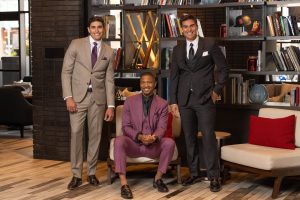 Three men pose indoors wearing formal suits in beige, burgundy, and black, standing and sitting in a modern lounge setting.