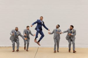 A groom jumps in the air while five groomsmen in gray suits reach toward him, laughing during a playful outdoor group photo.