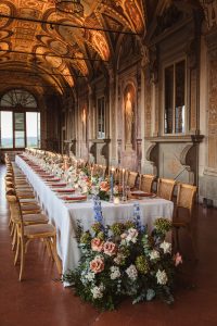 An elegant indoor wedding reception features a long table with candles, floral arrangements, and ornate painted walls.