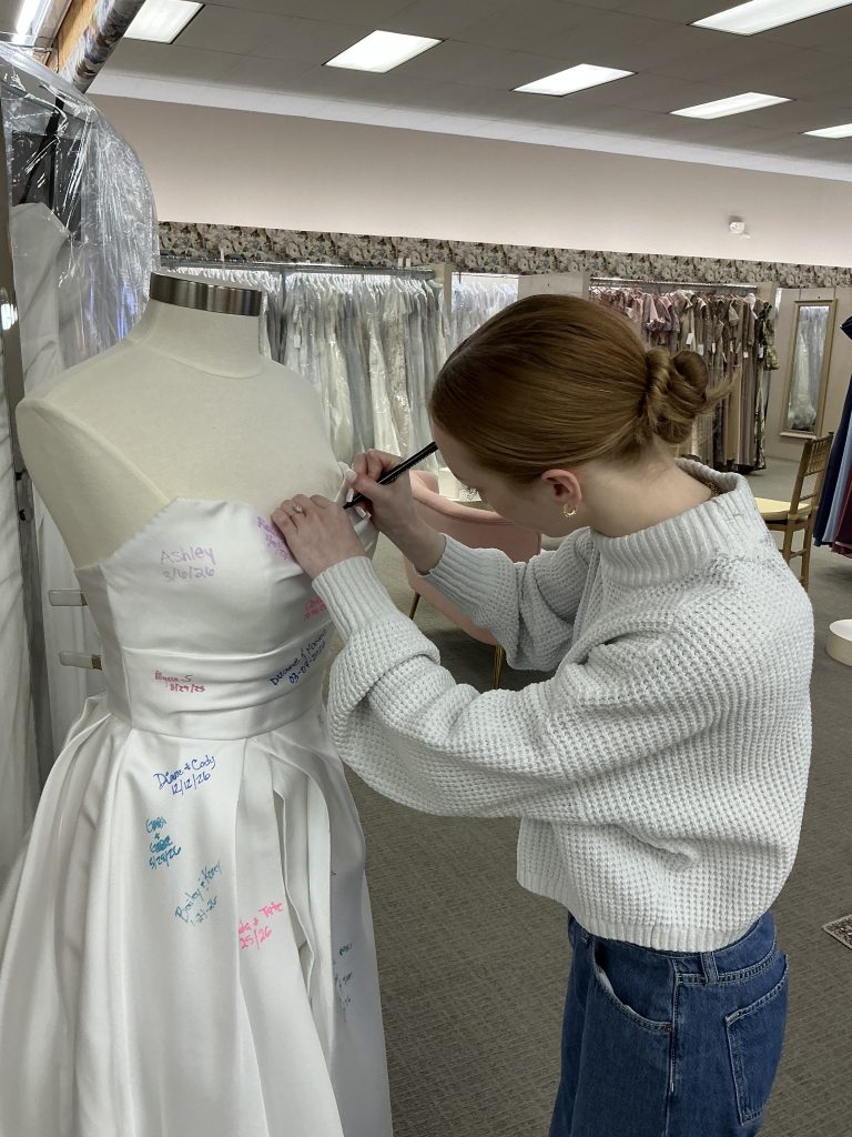A woman writes on a wedding dress displayed on a mannequin inside a bridal shop.