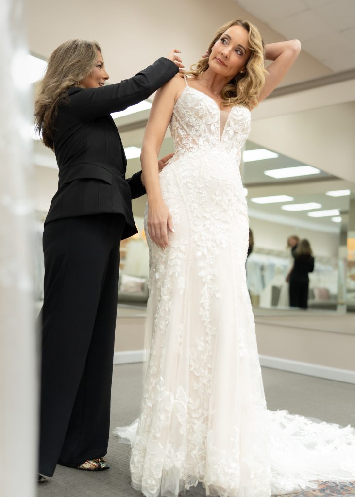 A bridal stylist adjusts the straps of a bride’s lace wedding dress during a fitting.