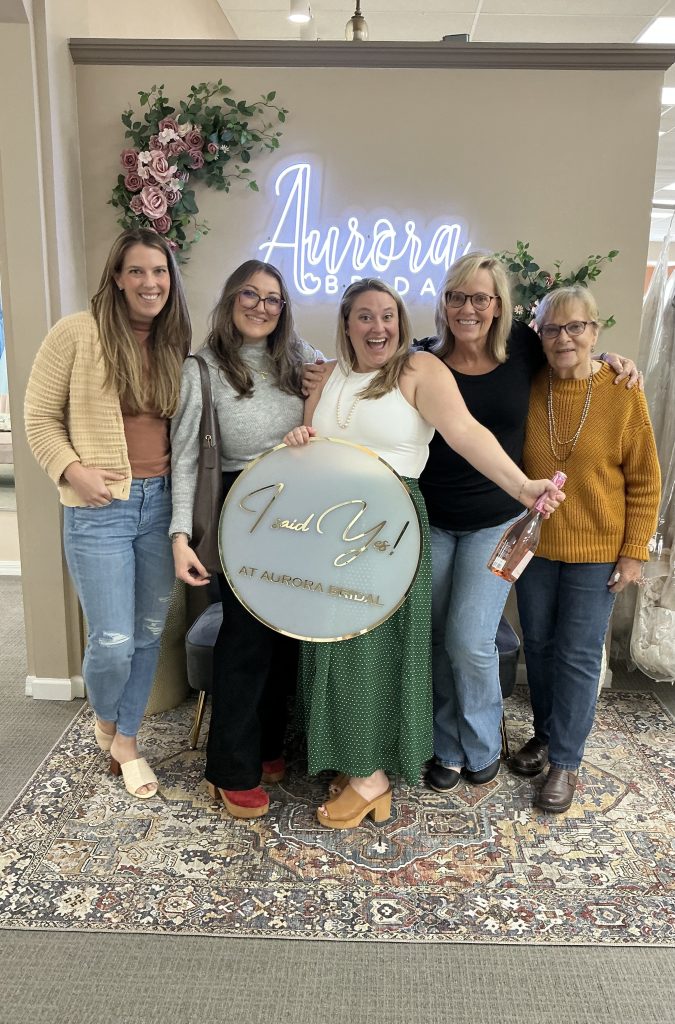 A group of women pose in front of a glowing “Aurora Bridal” sign while holding an “I said yes” sign and a bottle.