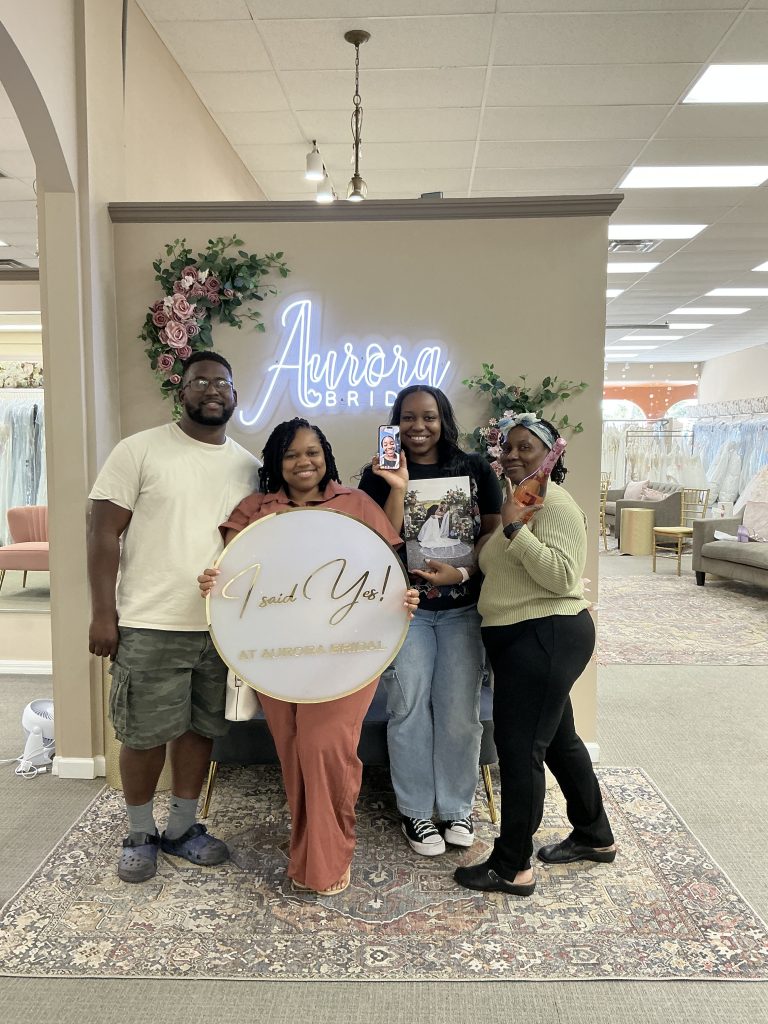 A group of four people pose in a bridal shop holding an “I said yes” sign in front of a lit wall logo.