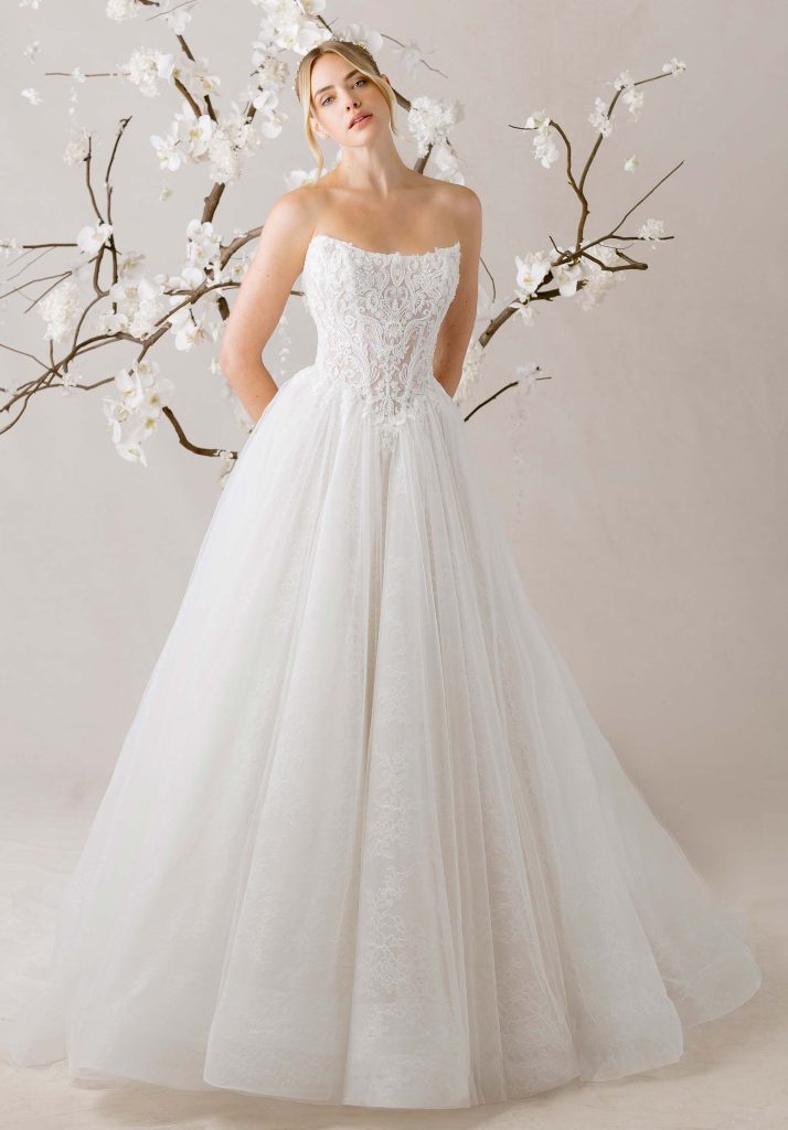 Bride in a strapless lace bodice ball gown posing in a studio with white floral branches behind her.