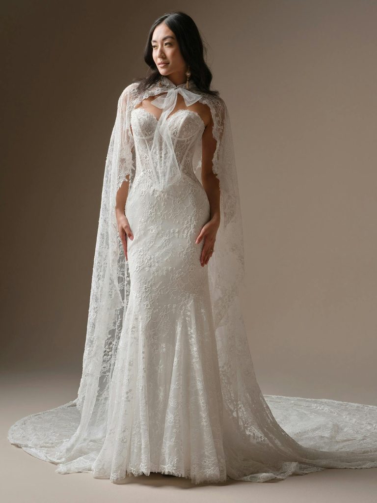 Bride wearing a fitted lace wedding gown with a sheer cape and bow detail standing against a neutral studio backdrop.