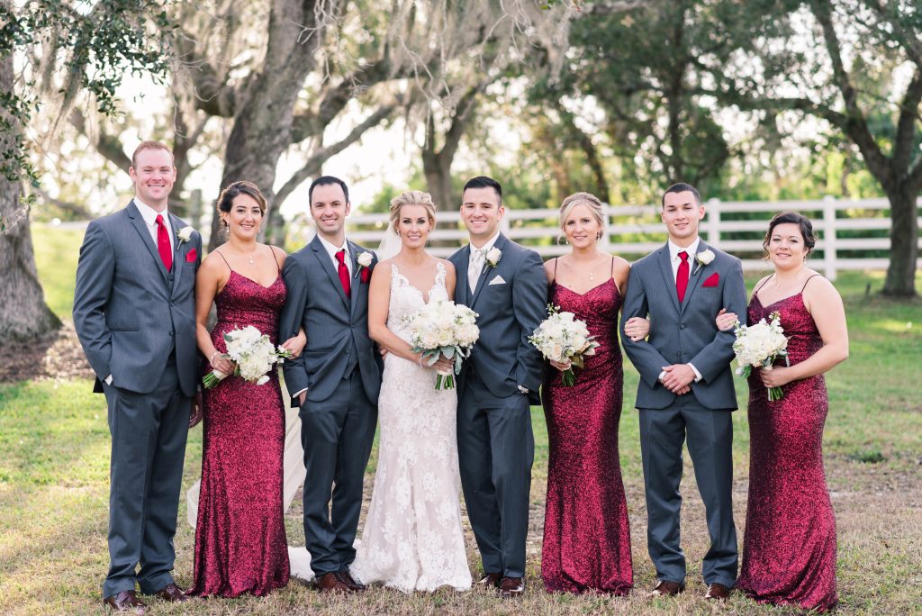 A bride and groom stand centered with their wedding party outdoors, featuring groomsmen in gray suits and bridesmaids in deep red dresses holding bouquets.