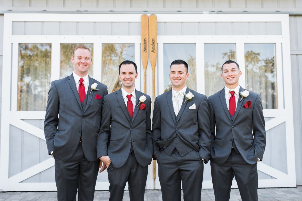 Four groomsmen wearing dark gray suits with red ties stand side by side in front of white barn-style doors.