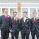 Four groomsmen wearing dark gray suits with red ties stand side by side in front of white barn-style doors.